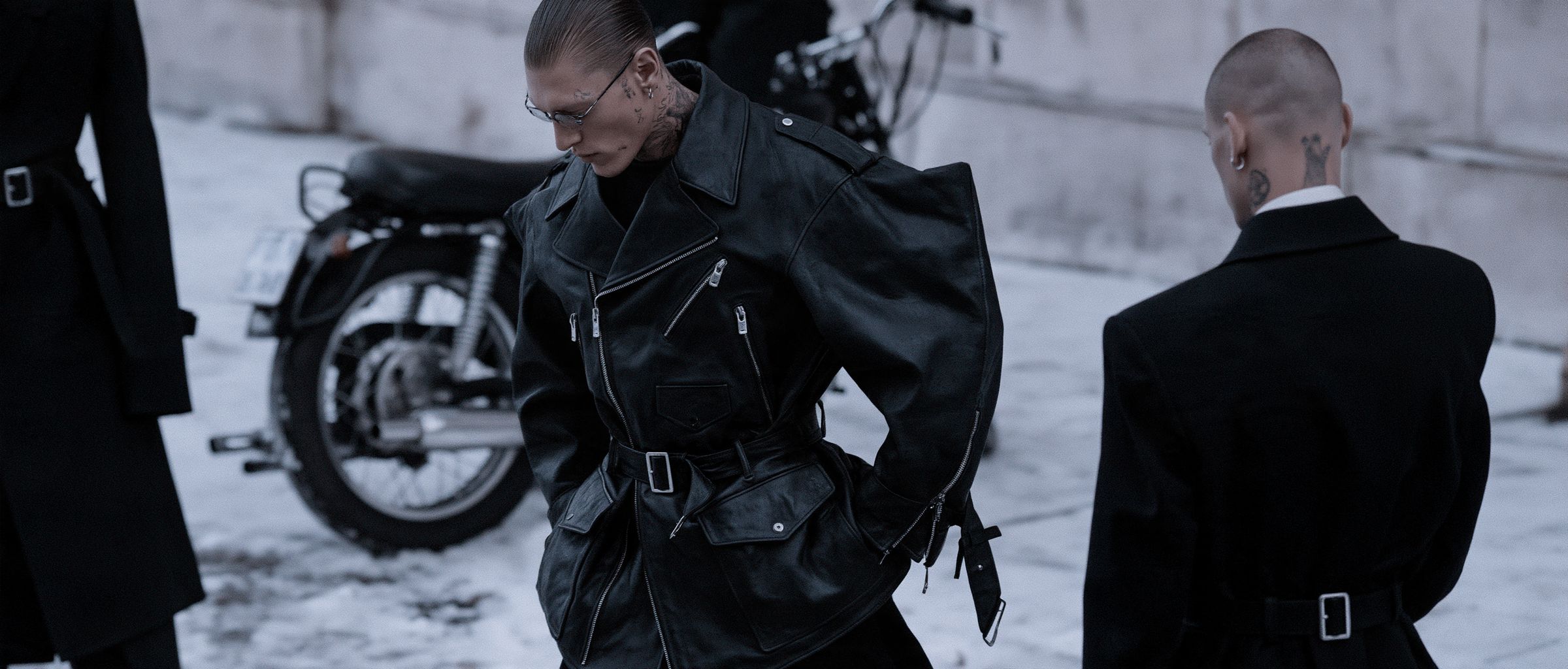 Three figures in black belted leather and wool coats beside a motorcycle on a snow-covered square